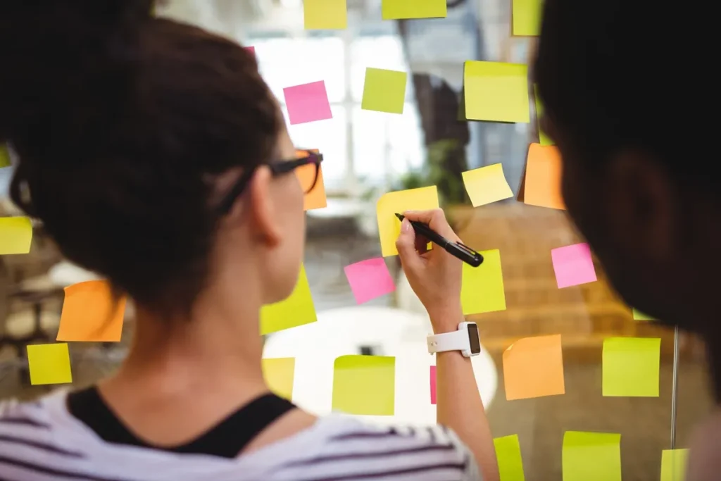 Mulher escrevendo em um post-it colado em um painel de vidro, organizando ideias para um planejamento estratégico.