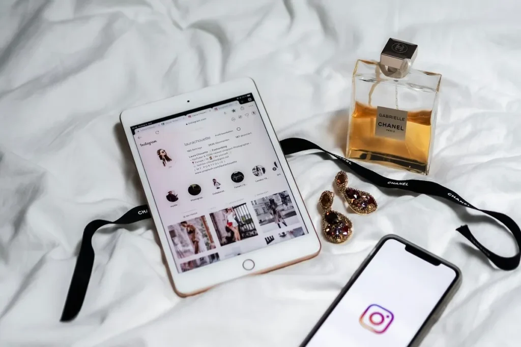 Tablet com perfil do Instagram exibido, ao lado de perfume Chanel, brincos e smartphone, sobre lençol branco.
