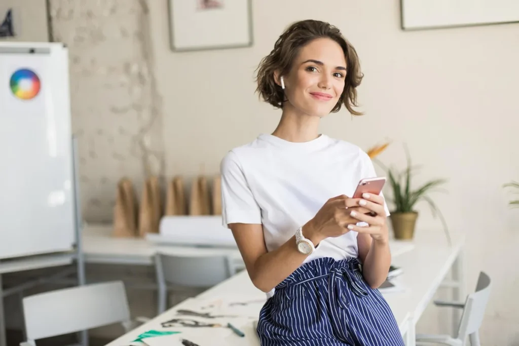 Mulher jovem sorridente usando fones de ouvido sem fio, segurando um smartphone, em ambiente de escritório moderno e criativo.