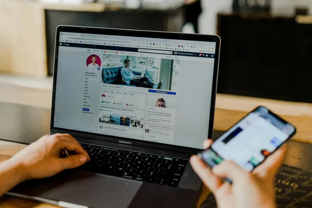 Pessoa utilizando um notebook MacBook Pro com o Facebook aberto, enquanto segura um smartphone, representando uso simultâneo de redes sociais para marketing digital ou gestão de conteúdo