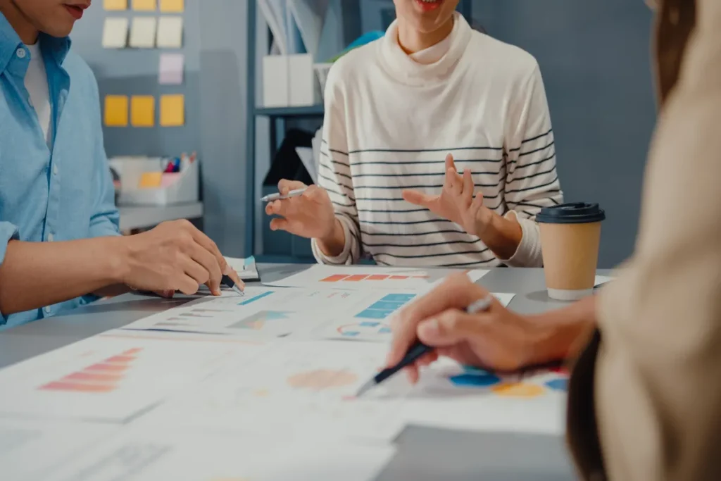 Profissionais reunidos ao redor de uma mesa analisando gráficos e documentos impressos, com canetas e café, durante uma reunião de planejamento estratégico ou briefing de marketing