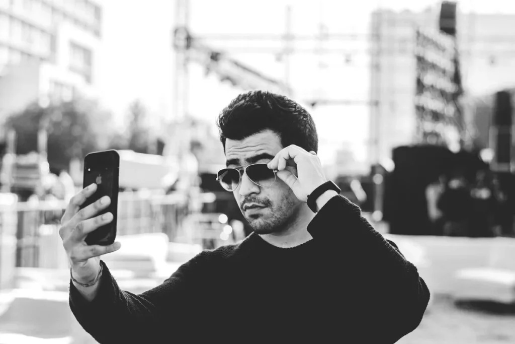 	Homem jovem usando óculos escuros tirando uma selfie com smartphone em ambiente externo