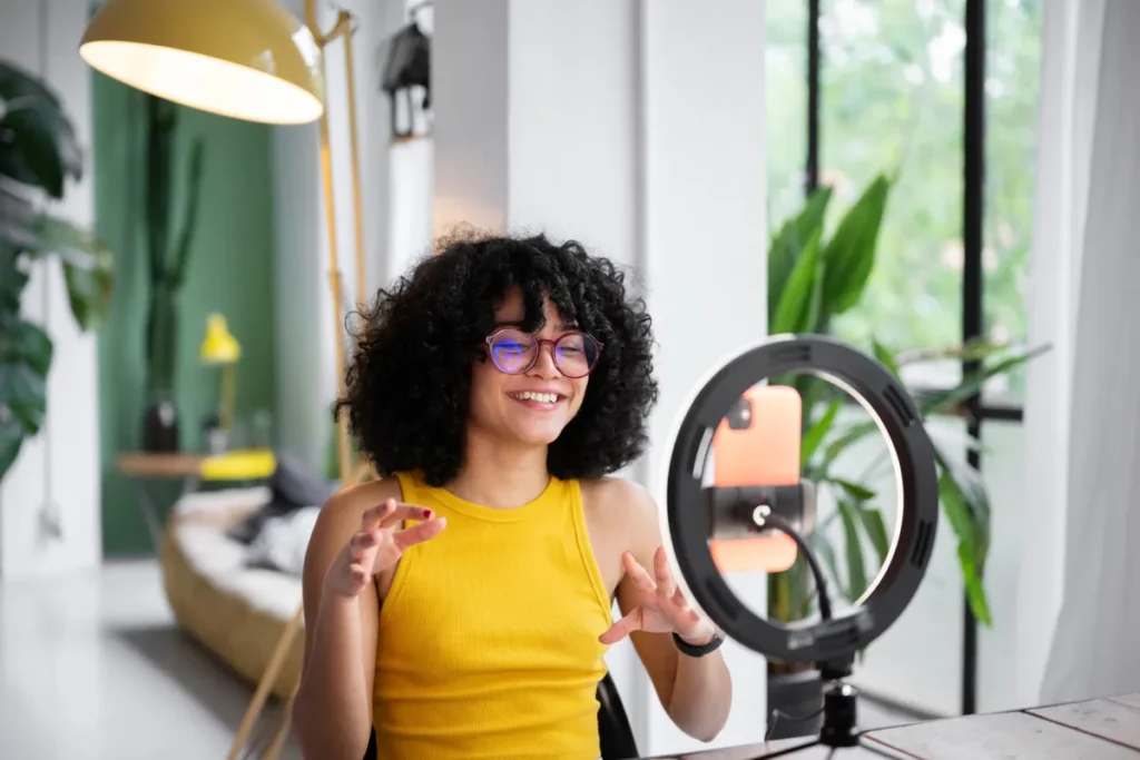 Mulher sorridente com cabelo cacheado gravando vídeo com smartphone e ring light em ambiente moderno