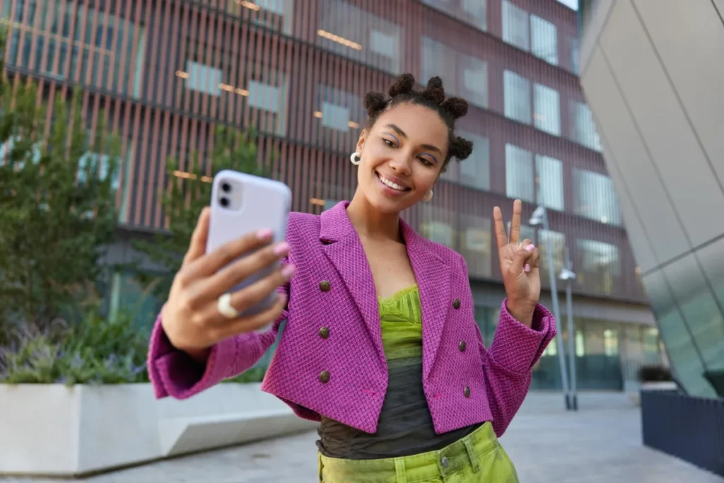 Mulher estilosa com cabelo preso em coques e roupa colorida tirando selfie com smartphone e fazendo sinal de paz em ambiente urbano moderno