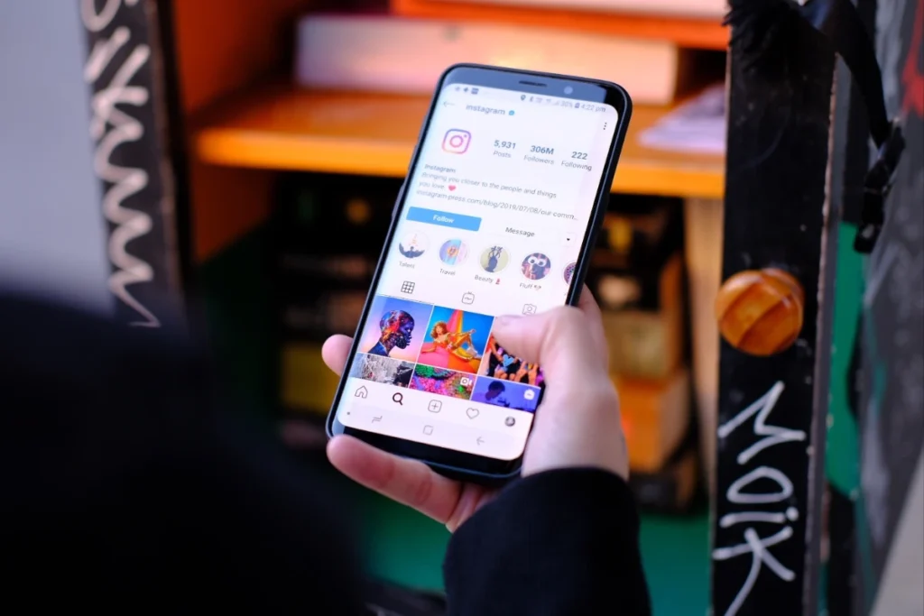 Mão segurando um smartphone onde se vê o perfil oficial do Instagram, com 5.931 posts, 306 M seguidores, botões de ‘Seguir’ e ‘Mensagem’, destaques circulares e grade de fotos coloridas