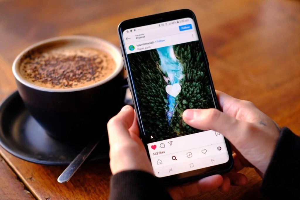 Mãos segurando um smartphone mostrando no Instagram uma foto aérea de um rio azul cortando uma floresta verde, com uma xícara de café ao lado em superfície de madeira