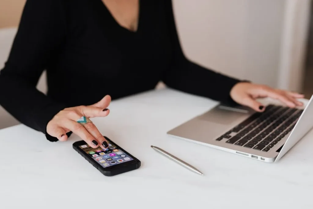 Mãos de uma pessoa utilizando um smartphone e um laptop em uma mesa branca, com aplicativos visíveis na tela do celular, representando a conectividade e o uso da tecnologia fundamental para empresas B2B.