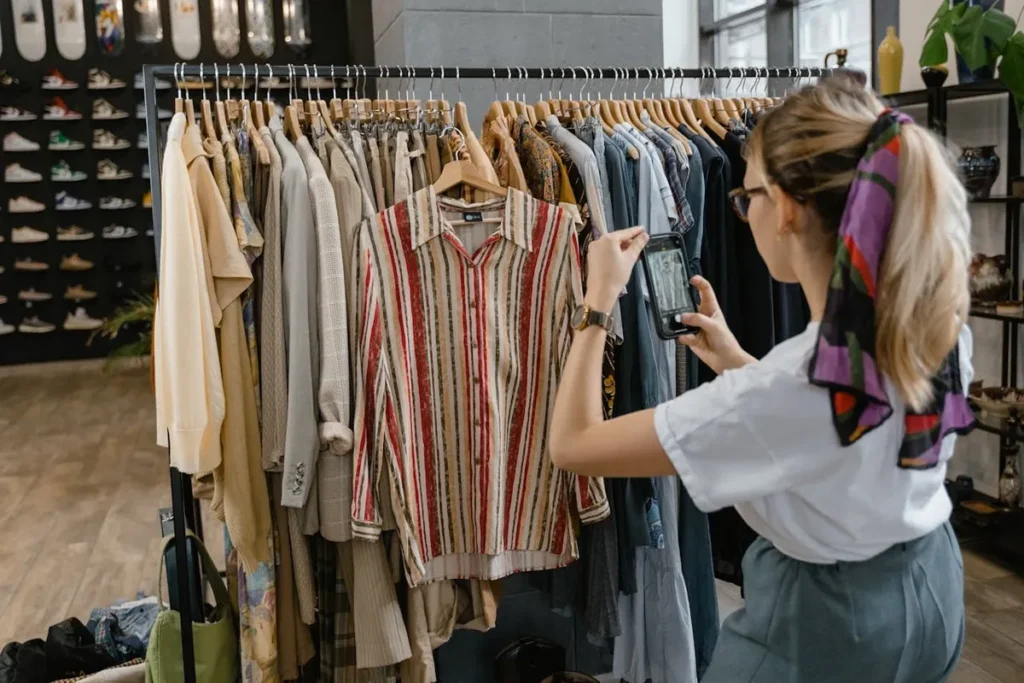 Mulher fotografando uma camisa listrada em um cabide de loja de roupas com seu smartphone, em um contexto que pode representar empresas B2B de atacado ou fornecimento de produtos para varejistas.