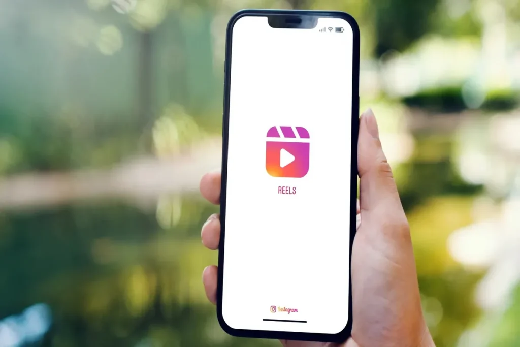 Uma mão segurando um smartphone com a tela exibindo o logotipo "Reels" do Instagram, com um fundo verde e desfocado de natureza. melhor horário para postar Reels