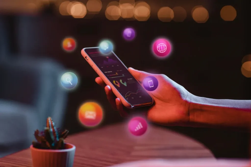 Mão de uma pessoa segurando um smartphone que exibe vários ícones flutuantes de aplicativos e redes sociais, representando a análise de métricas digitais.