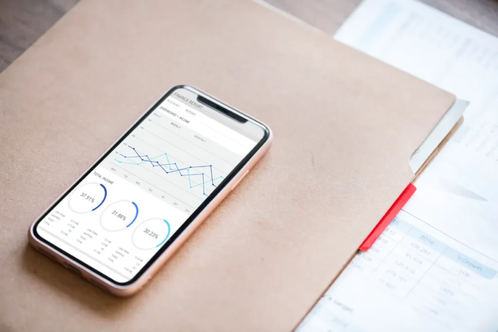 Smartphone exibindo um relatório financeiro com gráficos e porcentagens, sobre uma pasta e documentos, simbolizando a análise de métricas financeiras.