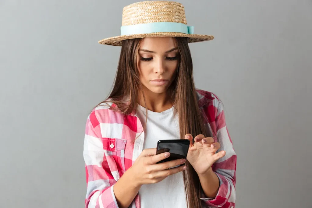 Mulher com chapéu de palha e camisa xadrez olhando para o celular, focada na tela, ilustrando a busca por informações ou o uso de palavras-chave para comunicação.