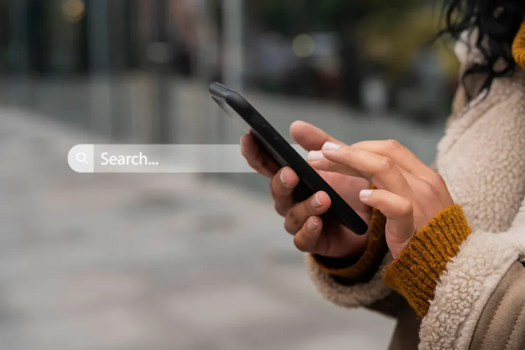 Mãos segurando smartphone com campo de busca 'Search...' visível, simbolizando a pesquisa de informações e a importância de palavras-chave para encontrar conteúdo.