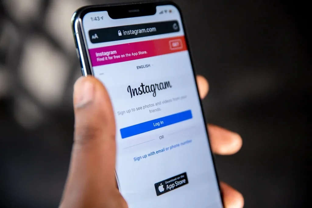 Mão segurando um smartphone com a tela exibindo a página de login do Instagram, com a opção de baixar o aplicativo na App Store.