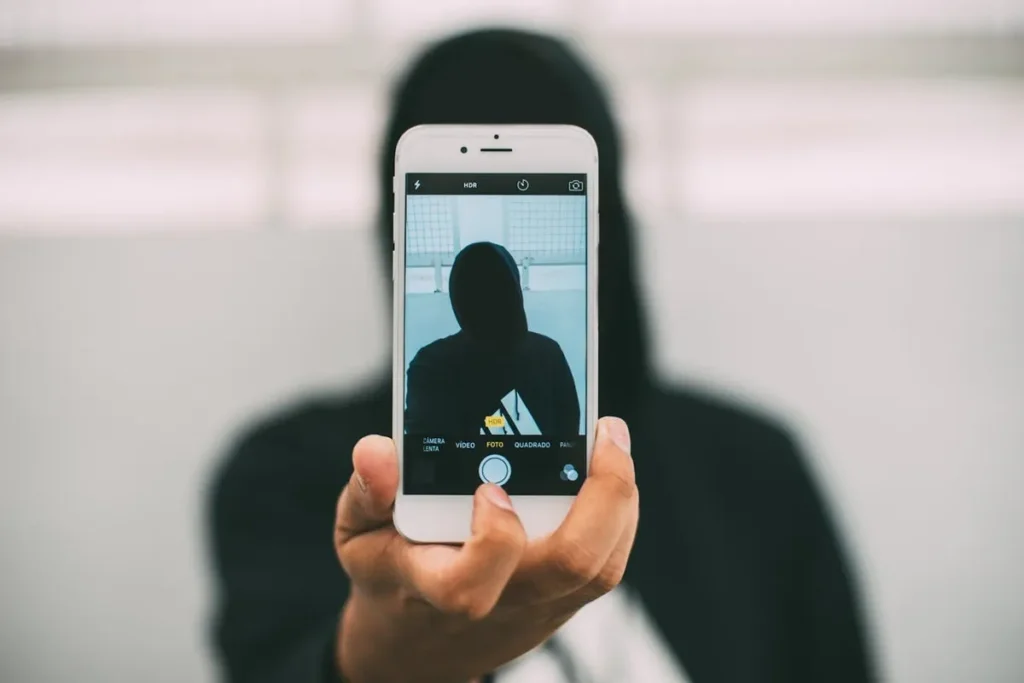Mão segurando um celular verticalmente, mostrando a tela da câmera frontal com uma silhueta escura de uma pessoa de capuz, ideal para ilustrar a criação de conteúdo misterioso ou anônimo para stories visando aumentar as visualizações.