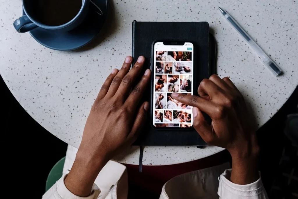 Visão aérea de mãos escuras sobre uma mesa clara, navegando em uma galeria de fotos de culinária ou café em um smartphone, com um caderno e caneta ao lado, sugerindo edição ou curadoria de conteúdo para stories.