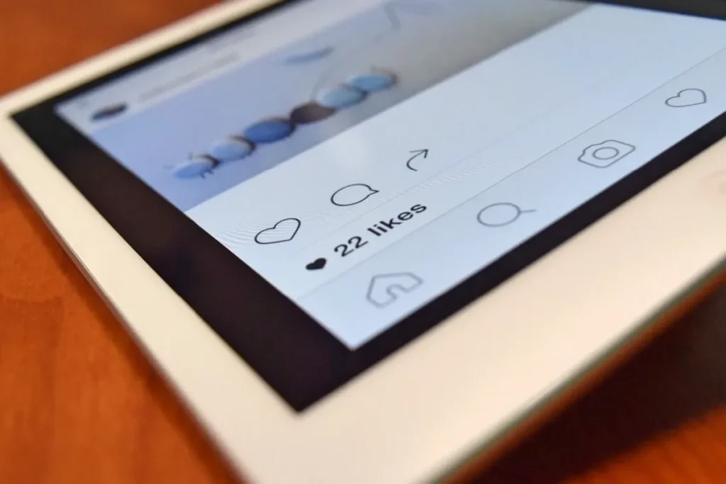 Close-up de um tablet exibindo uma interface de rede social com 22 curtidas, ícones de curtir, comentar, compartilhar, salvar, perfil e câmera, sobre uma superfície de madeira, simbolizando o engajamento e a visualização de conteúdo em plataformas digitais.
