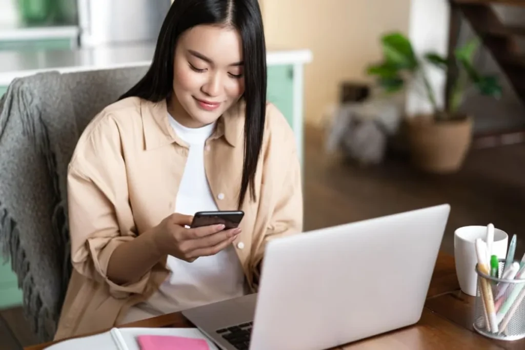 Mulher asiática sorrindo, usando o celular e o notebook, com a palavra-chave "Gerenciador de Anúncios Instagram".