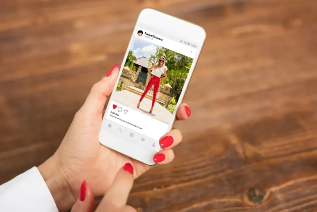 Mãos com unhas vermelhas segurando um celular com uma postagem de moda no Instagram, ilustrando o tom de voz e a identidade visual de uma marca.