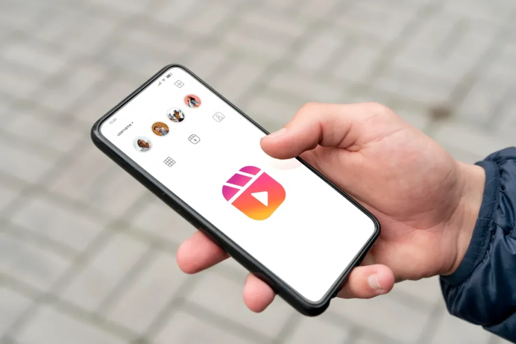 Mão segurando smartphone com o aplicativo do Instagram aberto, mostrando ícones de bio Instagram, destaques e o ícone do Reels em destaque.