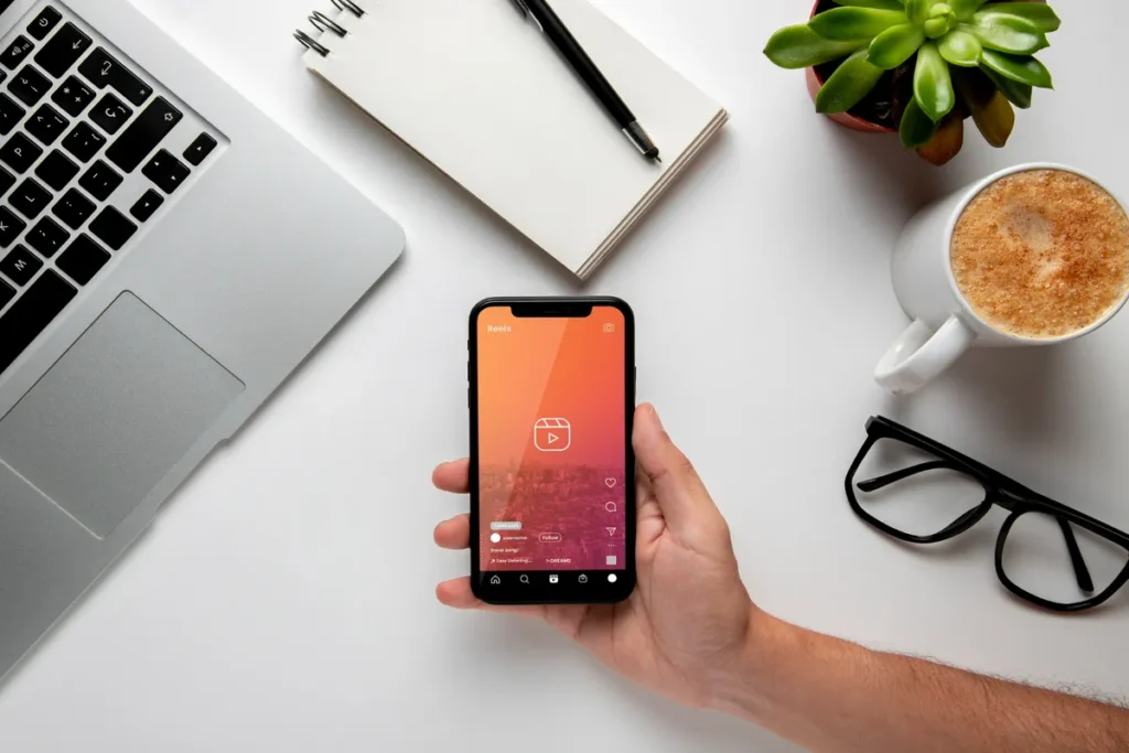 Mão segurando smartphone com o aplicativo do Instagram Reels aberto sobre uma mesa de escritório com notebook, caneta, agenda e café.