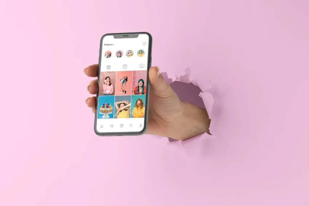 Uma mão saindo de um buraco em uma parede rosa, segurando um smartphone com um perfil do Instagram aberto, destacando a importância da bio Instagram e do feed.