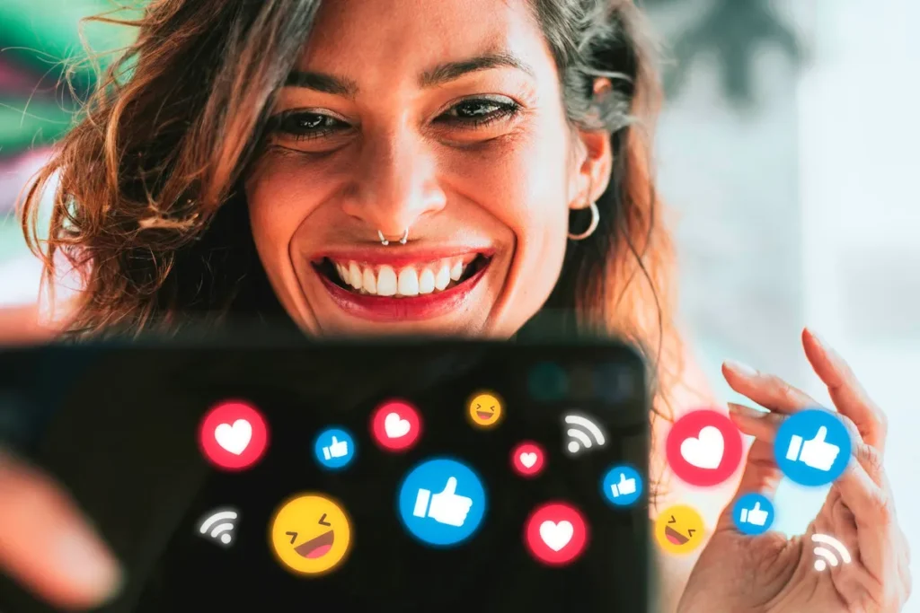 Mulher sorridente a fazer uma selfie com seu smartphone, cuja tela mostra ícones de curtidas (corações, polegares para cima) e emojis que indicam engajamento nas mídias sociais.