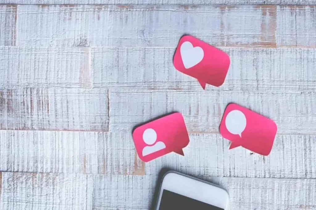 Três balões de fala cor-de-rosa, feitos de papel, com ícones de coração, perfil de pessoa e balão de fala, representando curtidas, comentários e seguidores em mídias sociais.