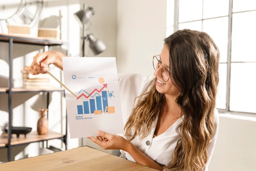 Mulher sorrindo, vestindo uma camisa branca e óculos, apontando com um lápis para um gráfico impresso que mostra barras azuis em crescimento e uma linha vermelha de tendência subindo, com a legenda 'COVID-19 Financial Recovery', simbolizando a análise de dados para Growth Marketing.