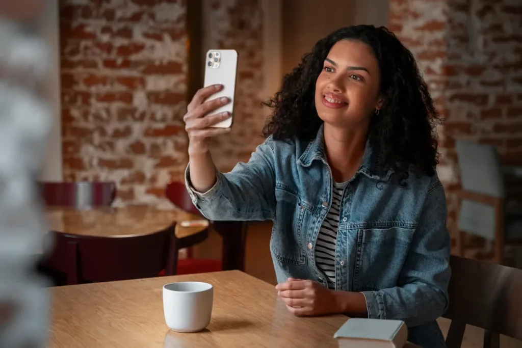 Retrato de uma mulher jovem, com cabelo cacheado e jaqueta jeans, sorrindo enquanto tira uma selfie ou faz uma videochamada com um smartphone em uma cafeteria rústica de tijolos aparente, simbolizando a presença digital e o marketing pessoal.