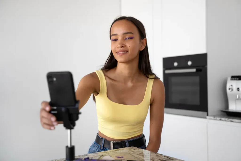 Uma jovem de pele morena clara, top amarelo e maquiagem vibrante, sorri enquanto grava um vídeo de si mesma com um smartphone em um tripé na cozinha, ilustrando a criação de conteúdo para marketing pessoal ou vlogs.