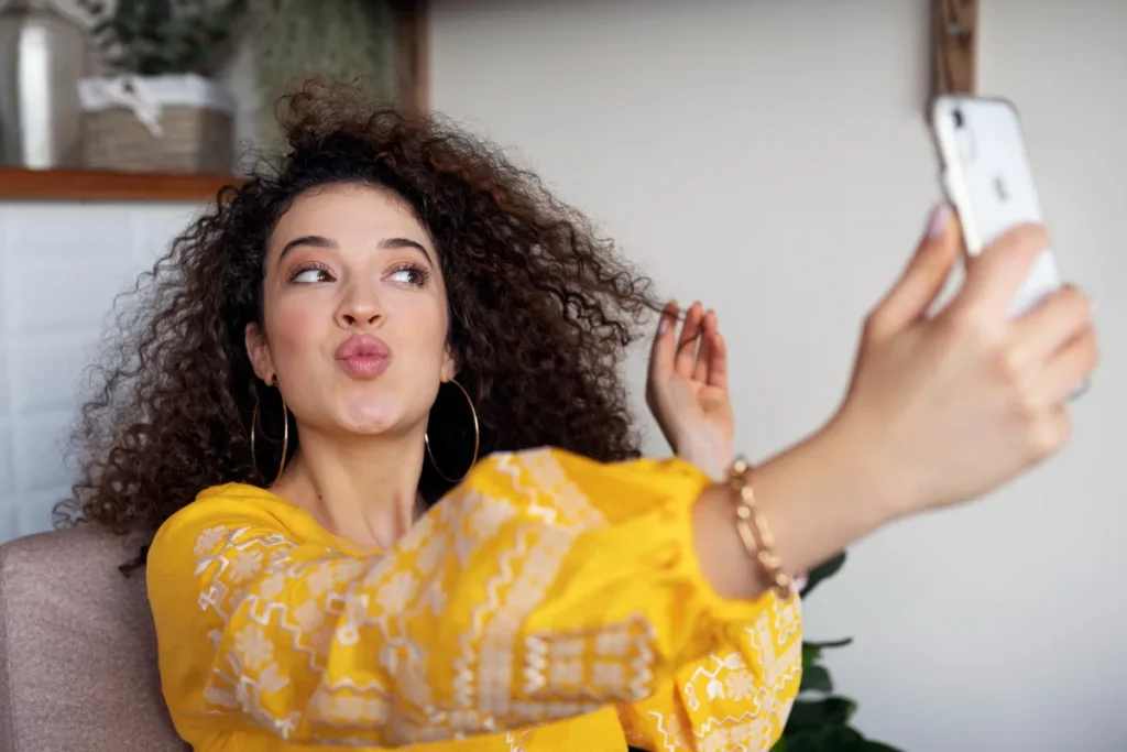 Retrato de uma mulher jovem, com longos cabelos cacheados e volumosos, vestindo uma blusa amarela estampada, fazendo uma "duck face" enquanto tira uma selfie com um smartphone, representando o marketing pessoal focado em beleza e lifestyle.