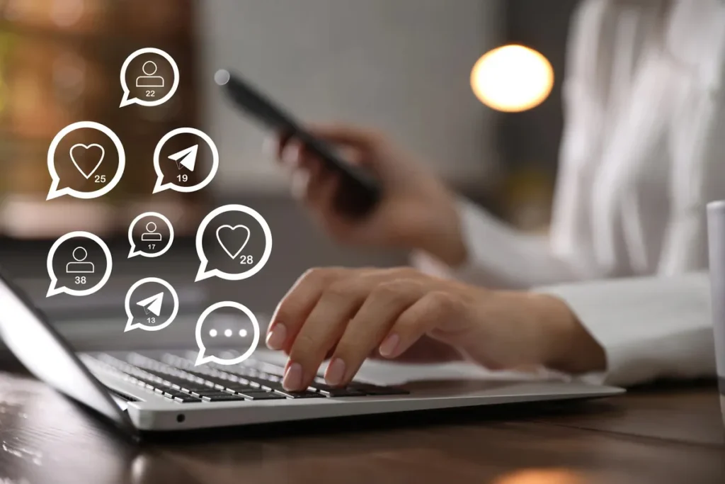 Pessoa digitando em um notebook enquanto ícones de redes sociais, como corações e balões de fala, flutuam sobre o teclado. Cena de monitoramento de mídias sociais e social listening.