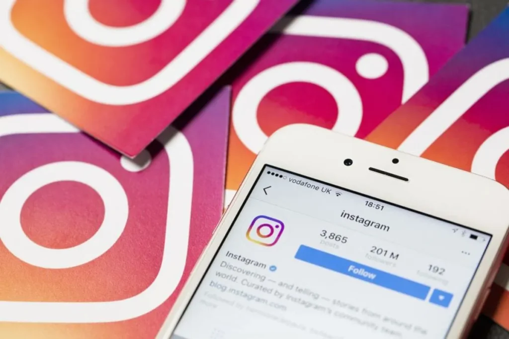Smartphone exibindo o perfil oficial do Instagram na tela, com a contagem de seguidores e posts, sobrepondo vários logotipos impressos do Instagram em fundo degradê.