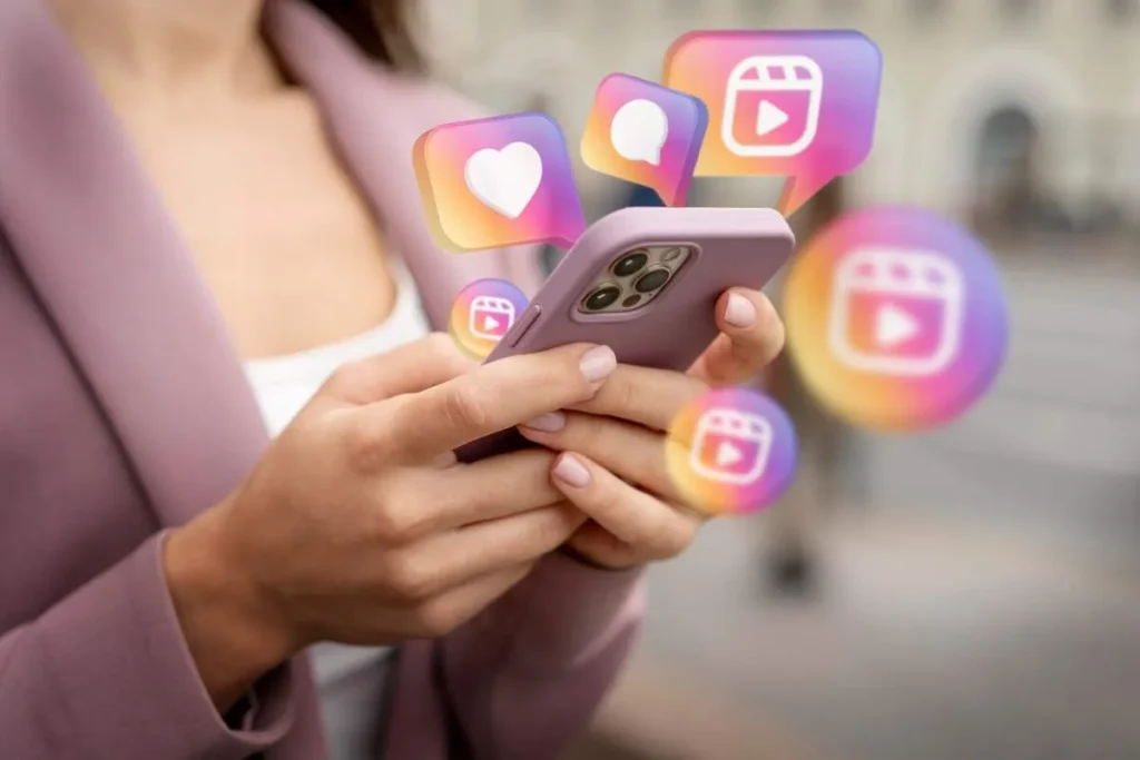 Pessoa segurando um celular na vertical com ícones 3D flutuantes do Instagram ao redor, como coração (like), balão de diálogo (comentário) e o símbolo do Reels, representando a interação com a plataforma e o algoritmo.
