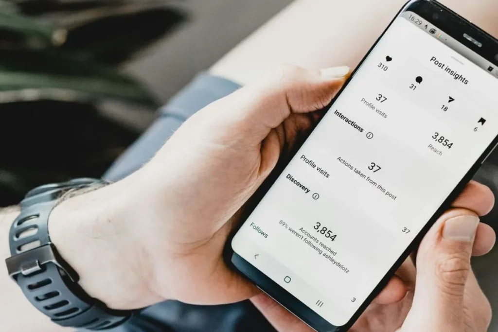 Uma pessoa segurando um smartphone que exibe a tela de Insights (estatísticas) de um post do Instagram, mostrando métricas como alcance (Reach), visitas ao perfil, interações e descoberta.