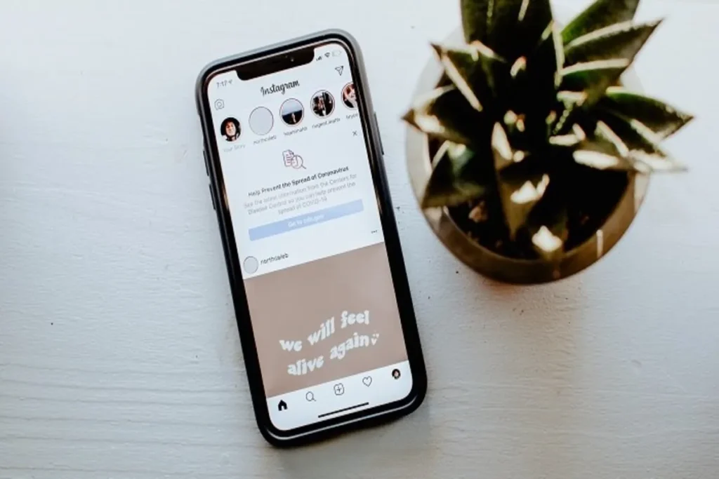 Smartphone exibindo o feed do Instagram sobre uma superfície branca e simples, com um post sobre esperança ("we will feel alive again") e um aviso de prevenção ao coronavírus, ao lado de um vaso de planta verde.