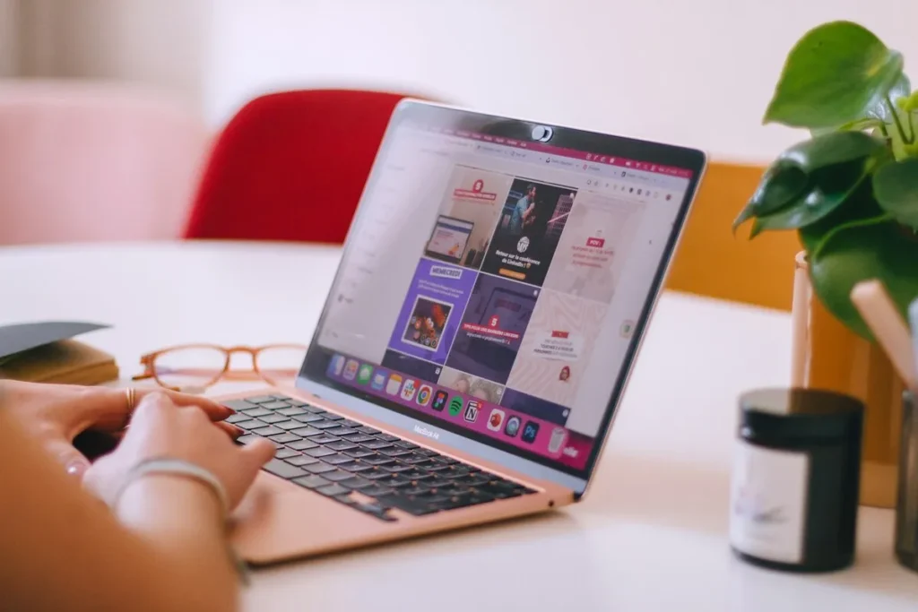 Close-up de uma mulher usando um MacBook Air com a tela aberta em um dashboard ou plataforma de gerenciamento de conteúdo visual para mídias sociais (Pinterest, Instagram, etc.), com óculos e itens de papelaria na mesa.